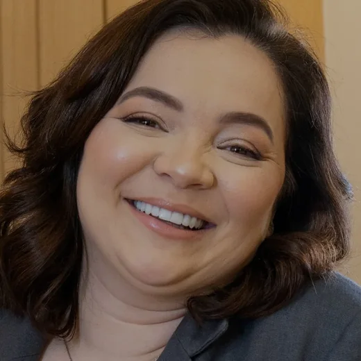 Dra. Beatriz Alves rindo, de blazer cinza, diante do painel de carvalho