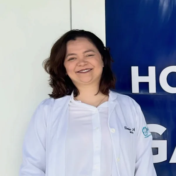 Dra. Beatriz Alves de jaleco branco, sorrindo, diante de uma fachada azul de hospital